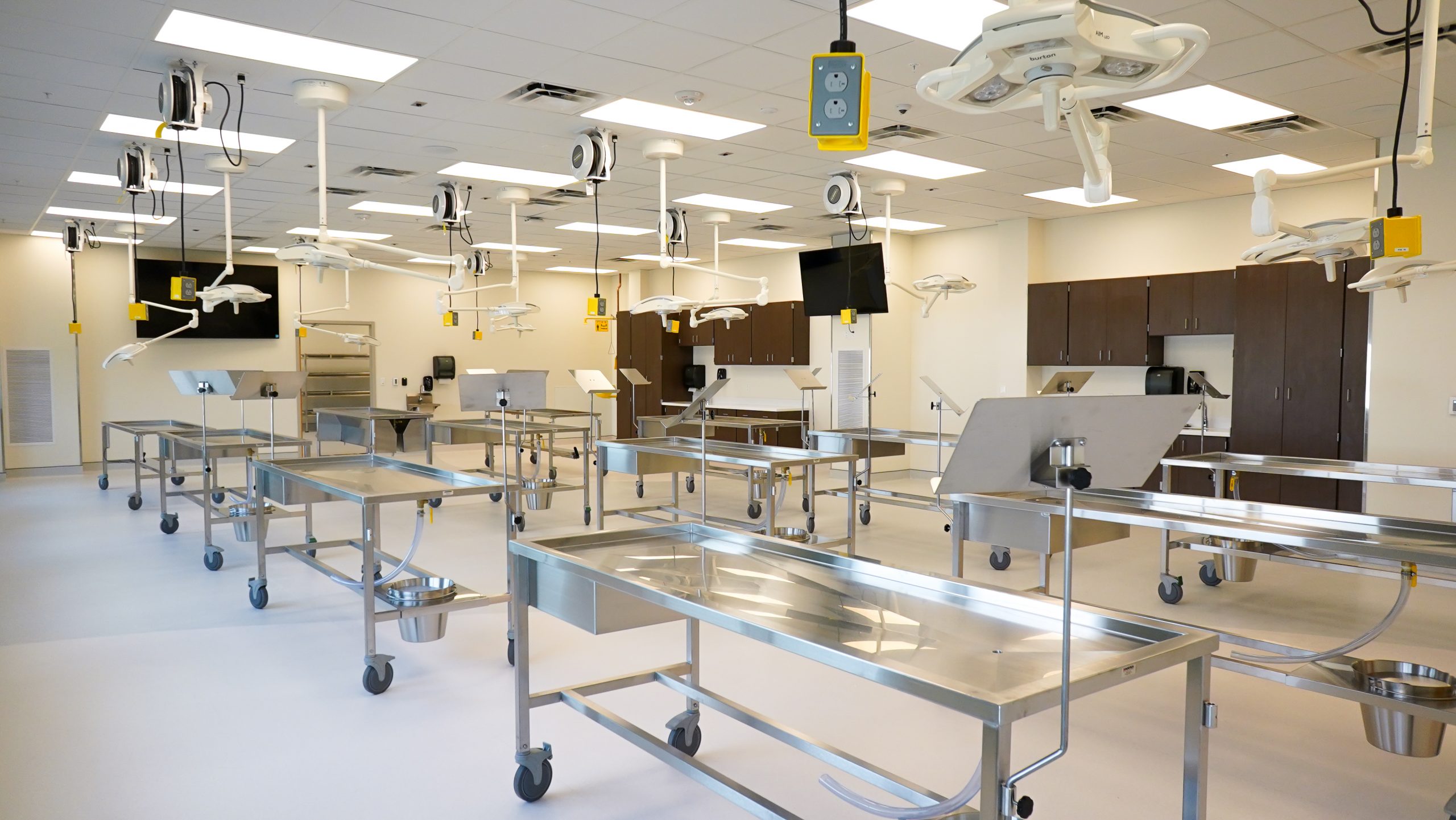 Large anatomy laboratory with multiple stainless steel dissection tables on wheels, overhead surgical lights, monitors, and ceiling-mounted electrical outlets. The space has neutral walls and built-in cabinetry.
