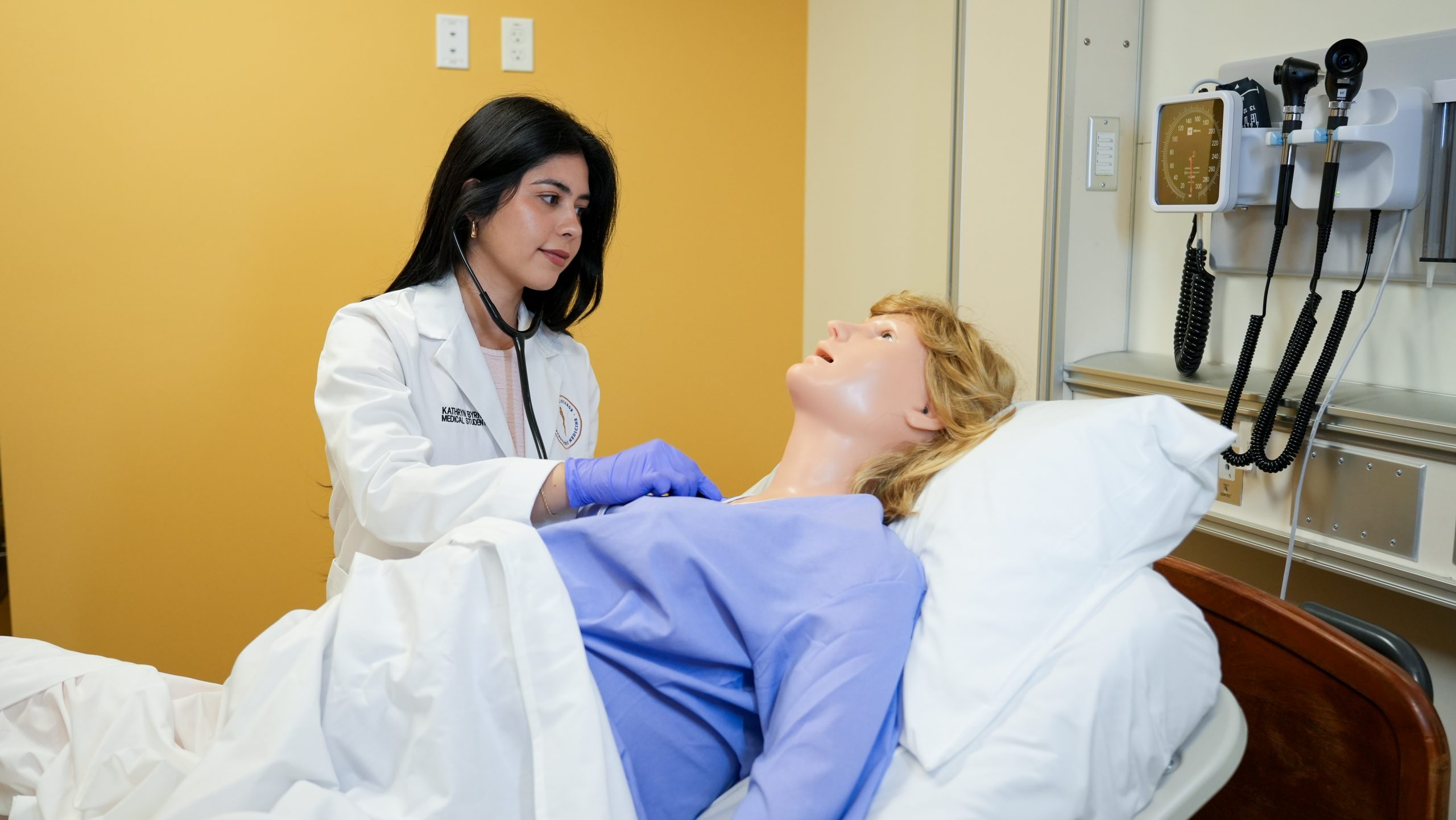 A medical student holds a stethoscope up to a medical mannequin.