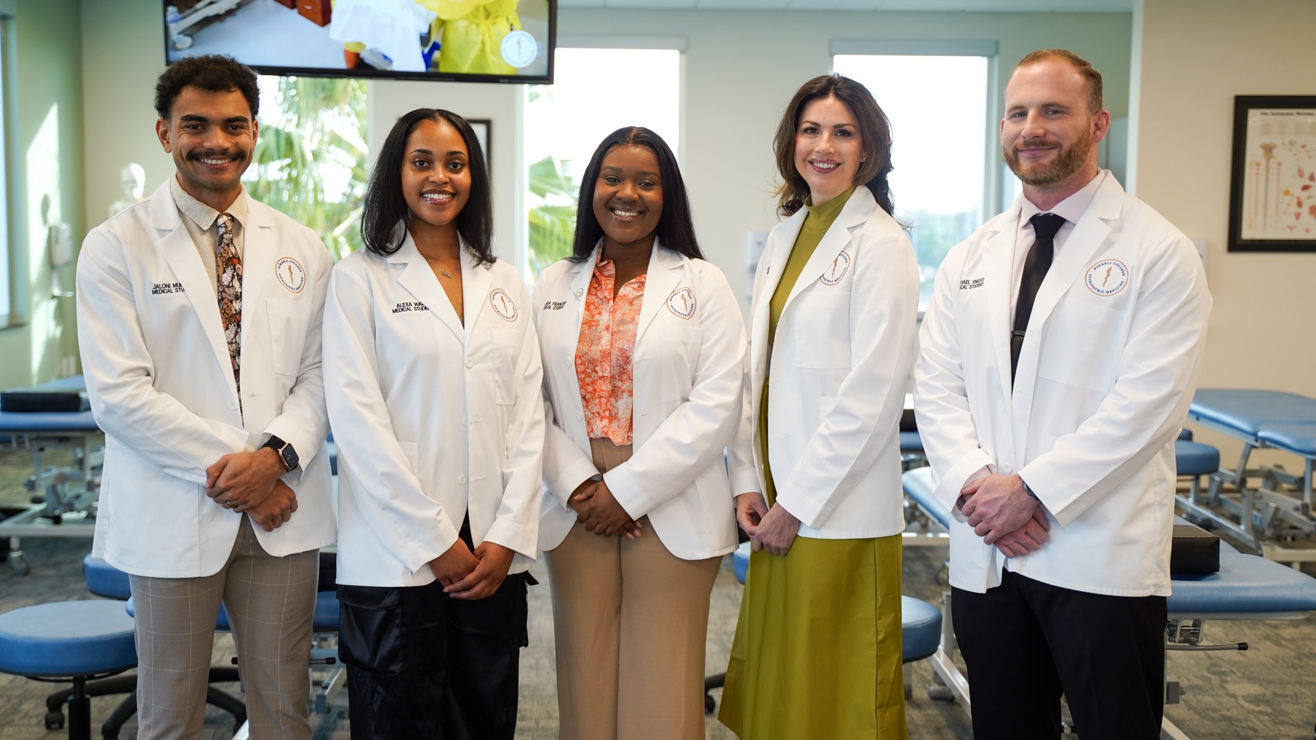 A group of medical students in white lab coats smile with their hands folded while standing in an exam classroom.