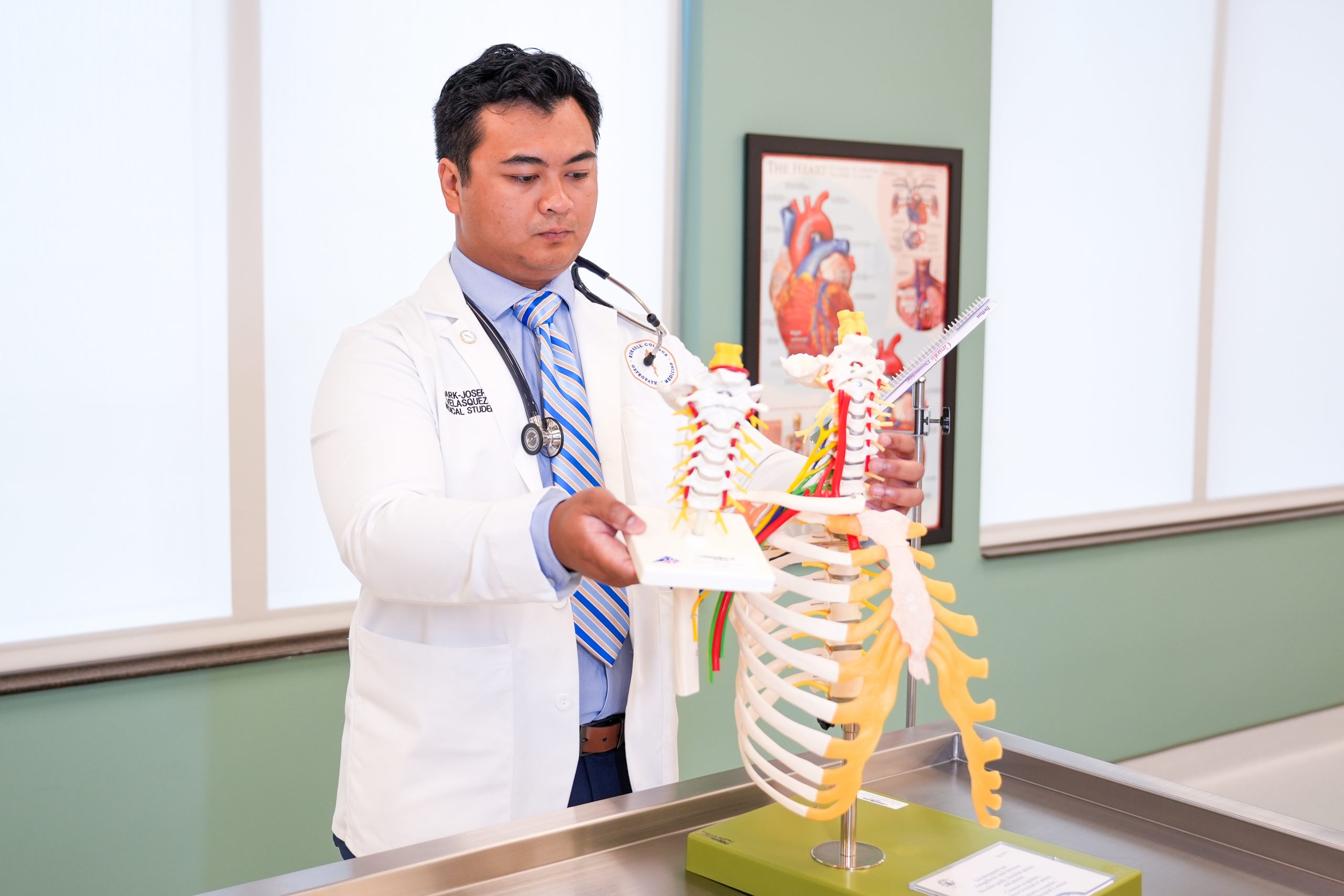 A medical student examines a model of the spine in a classroom, with a heart anatomy poster visible in the background.