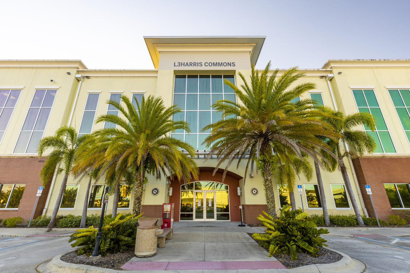 Exterior view of the L3Harris Commons building, featuring large windows, palm trees, and landscaped greenery at the entrance.