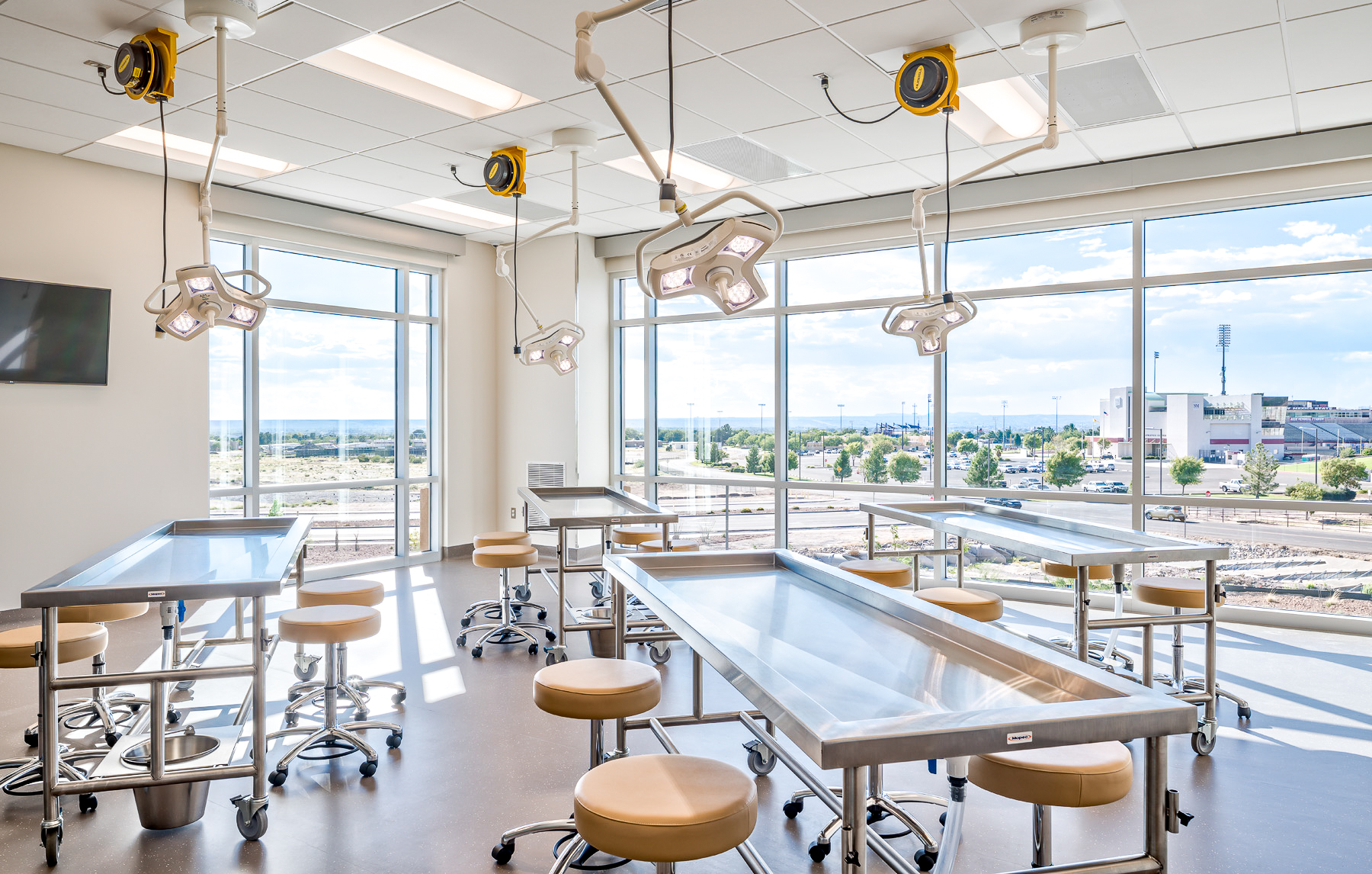 Anatomy lab with stainless steel dissection tables, stools, and overhead surgical lights. Large windows provide natural light and overlook a campus and stadium outside.