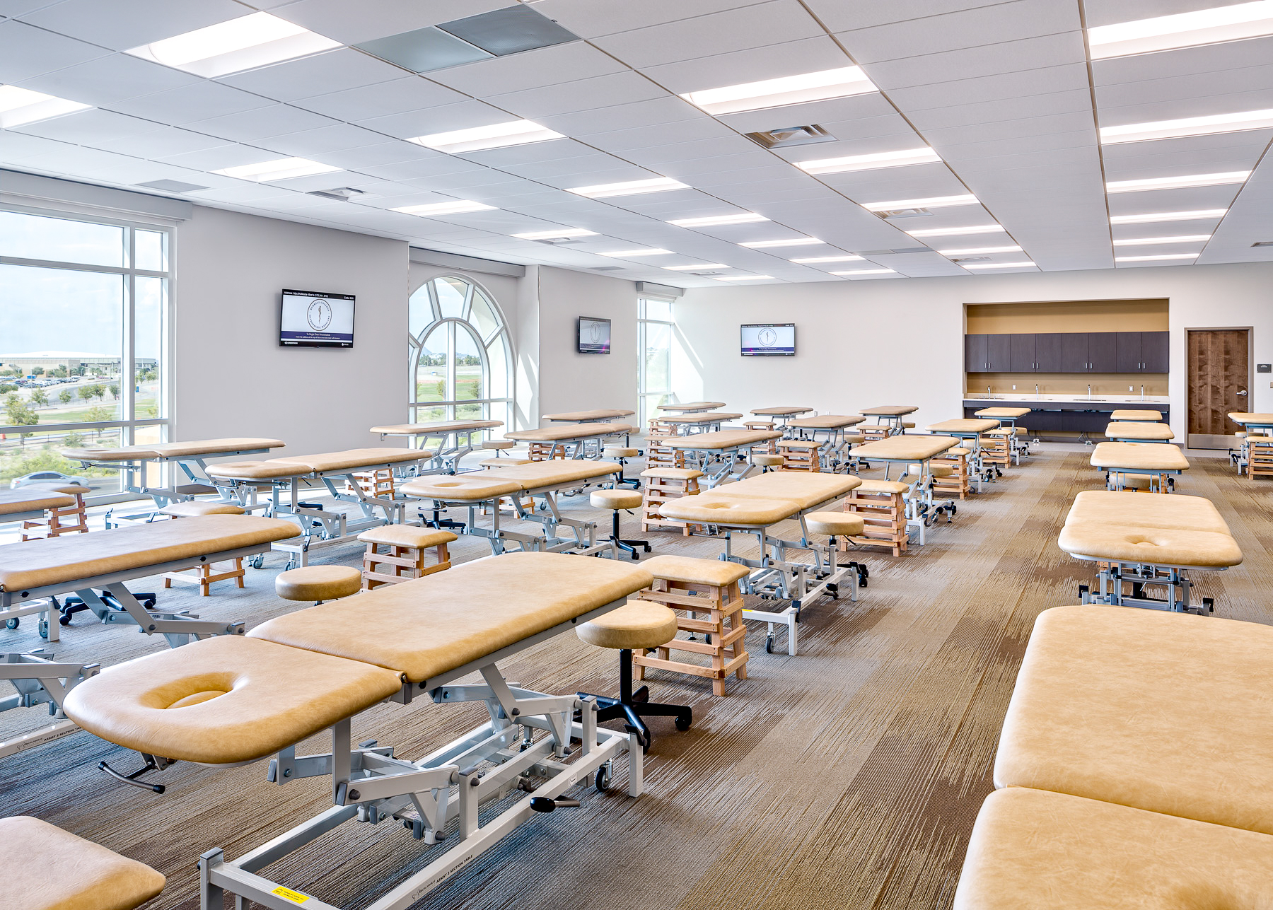 Spacious osteopathic manipulative medicine (OMM) lab with rows of padded examination tables, rolling stools, and large windows. Wall-mounted monitors and cabinetry are visible in the background.