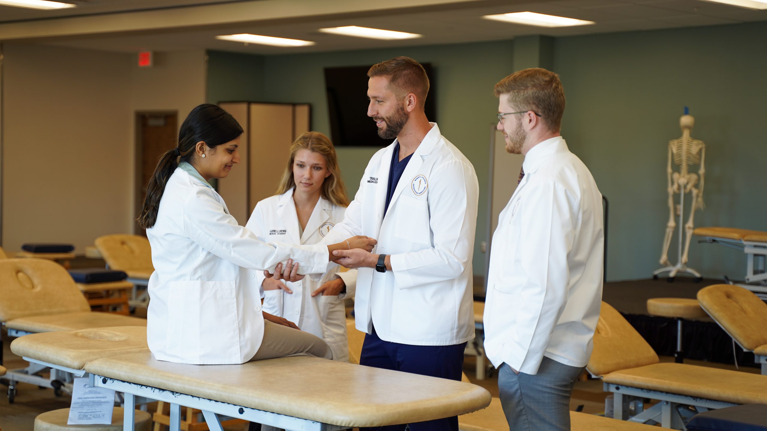 In a classroom setting, a medical student demonstrates an arm examination on a classmate while other students watch.