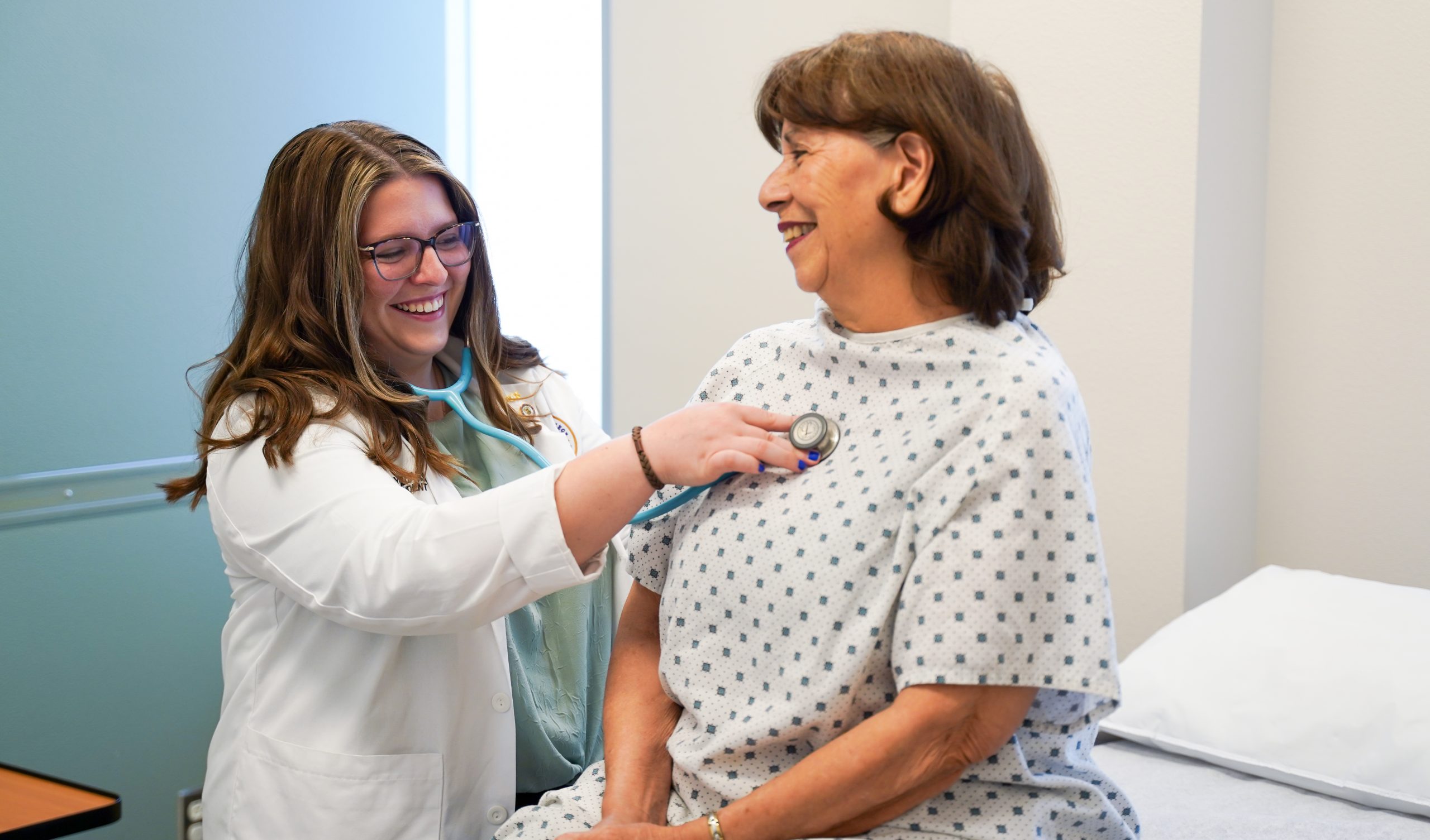 A medical student holding a stethoscope up to a patient's chest while they smile at each other.
